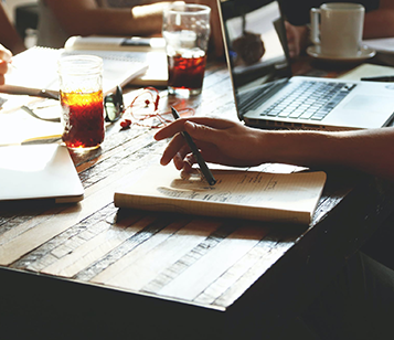 A man is holding a pen in his hand, and there are notebooks , Laptop , drinks & coffee cups and documents on the table